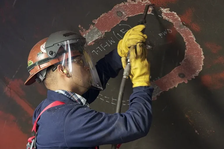 Ship hull spot repair vs full recoat: shipyard worker chipping old paint from a ship hull